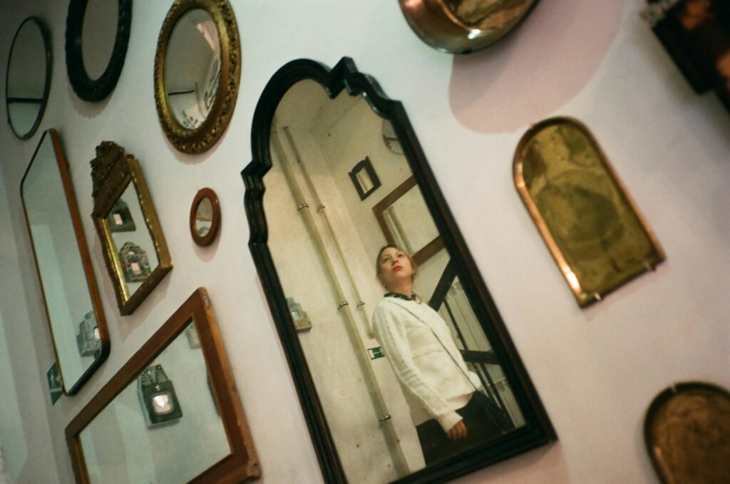 Contemplative young female in casual wear standing on stairs and reflecting in one of many framed mirrors hanging on wall